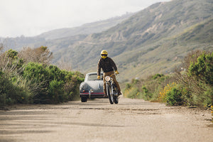 Motorbike racing ahead of the Porsche through mountainous landscape. 